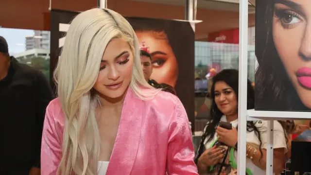 Business Boss- Kylie Jenner at a Kylie Cosmetics launch event, holding lip kits and posing in front of the brand logo.